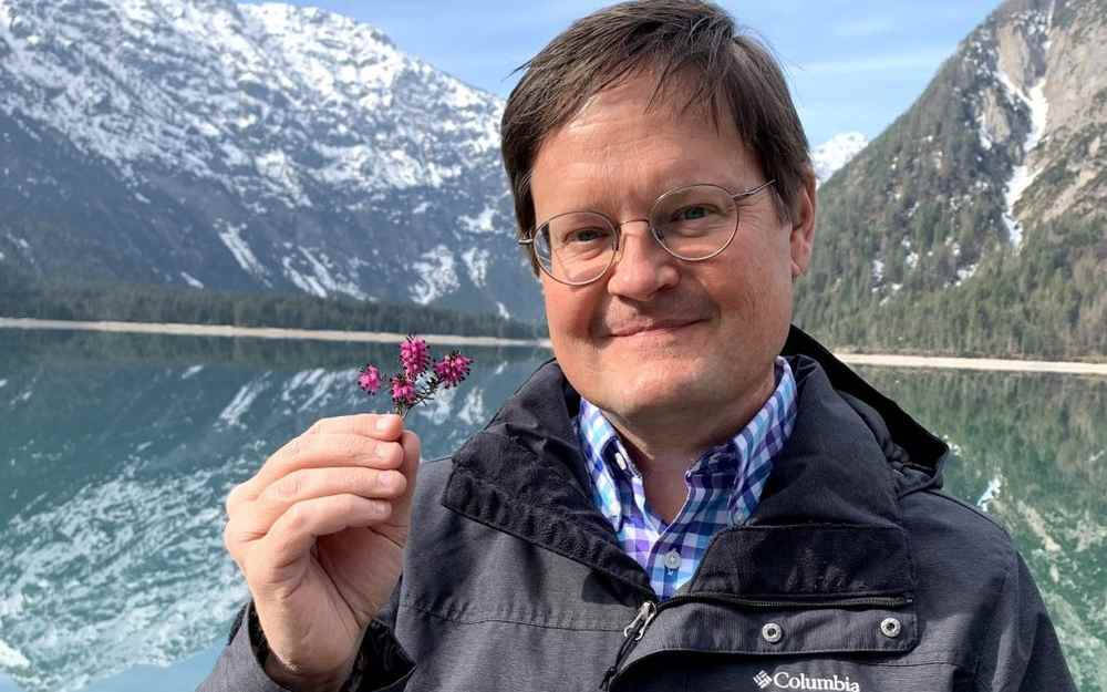 A man wearing glasses and a dark Columbia jacket smiles while holding a small purple flower in front of a scenic lake surrounded by snow-capped mountains and pine trees. The clear blue sky and reflections on the water suggest a calm, cool day in a mountai